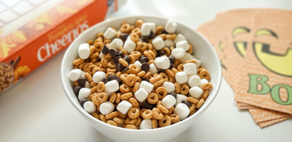 white bowl of cheerios, marshmallows and chocolate chips with cheerios cereal box and halloween theme goody bags in the background