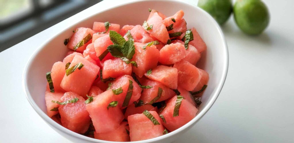 bowl of watermelon cubes with mint strips. Two limes in background.