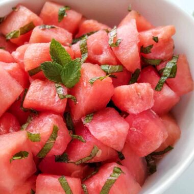 bowl of cut watermelon cubes mixed with strips of mint leaves. Whole limes in the background.