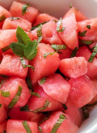 bowl of cut watermelon cubes mixed with strips of mint leaves. Whole limes in the background.