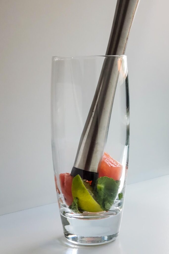 tall cocktail glass with lime mint and watermelon cubes at bottom and a muddler sticking out of it