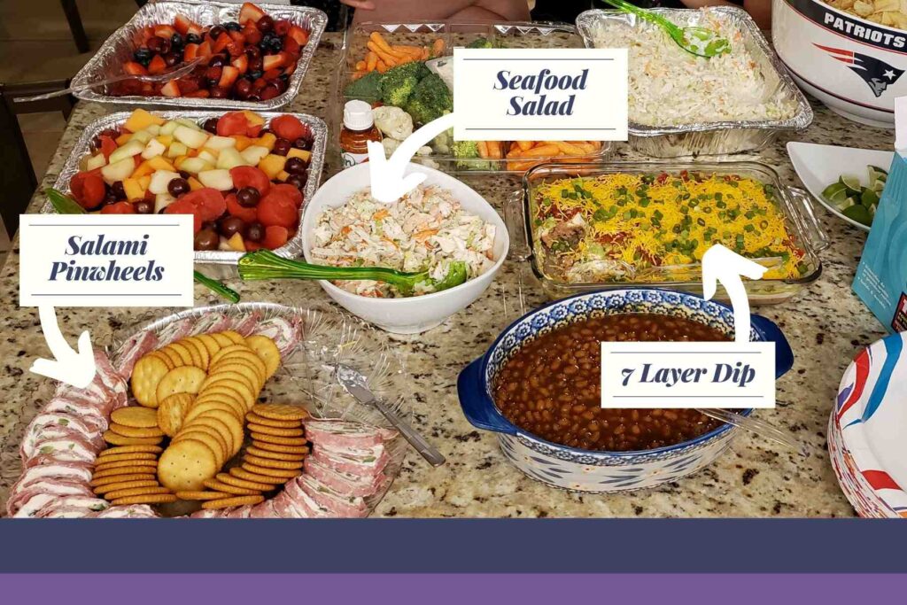 Variety of foods on counter highlighting the salami cream cheese rollups on a dish with ritz crackers; the seafood salad in a white bowl with green spoon; and the 7 layer dip in a glass casserole dip