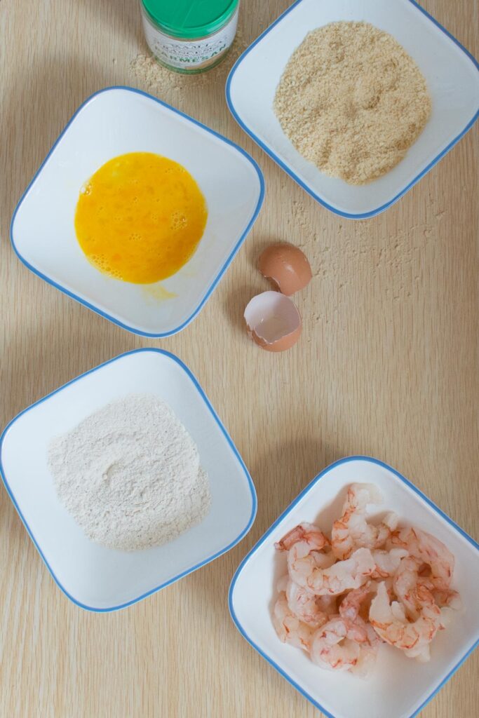 4 white bowls holding the ingredients for breading the shrimp; one with parmesan and breadcrumbs, one with beaten egg, one with flour, and one with red argentinian shrimp. a cracked eggshell is on the table too