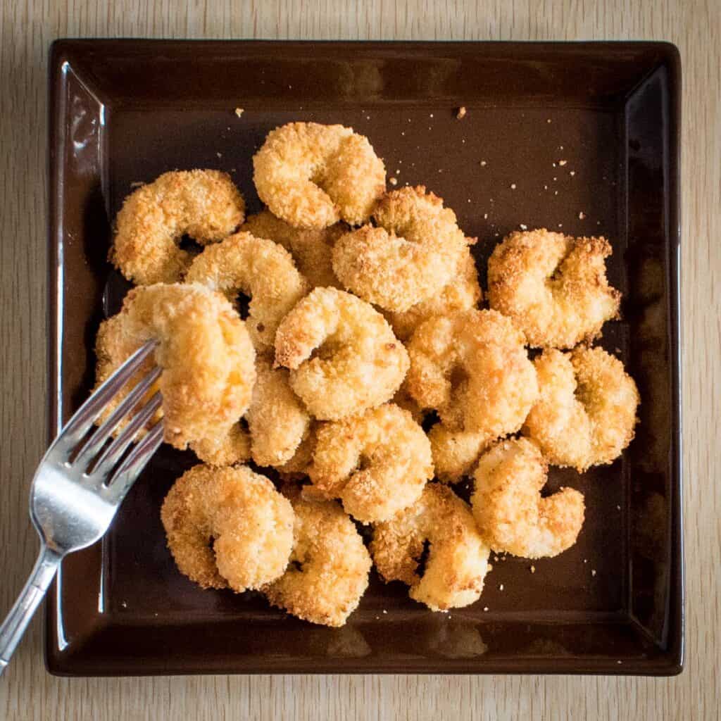 pile of breaded baked shrimp on a brown square plate with a silver metal fork holding up one shrimp
