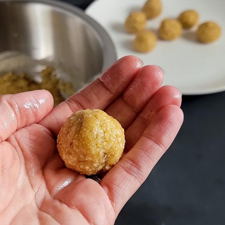 palm of hand holding one rolled matzo ball about the size of a walnut. Bowl of mix and plate of rolled balls in the background.