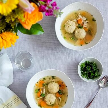 Two white bowls with matzo ball soup. Bouquet of flowers in top left corner. Glass of water. Small bowl of parsley garnish. White and yellow linen napkin and a spoon.