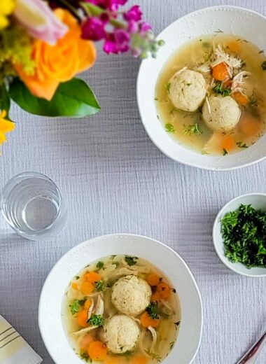 Two white bowls with matzo ball soup. Bouquet of flowers in top left corner. Glass of water. Small bowl of parsley garnish. White and yellow linen napkin and a spoon.