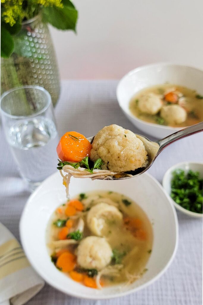 Close up of a spoon with a bite of matzo ball soup with carrot, parsley and chicken. Background of table with flower vase, two bowls of soup, parsley garnish, and a cup of water.