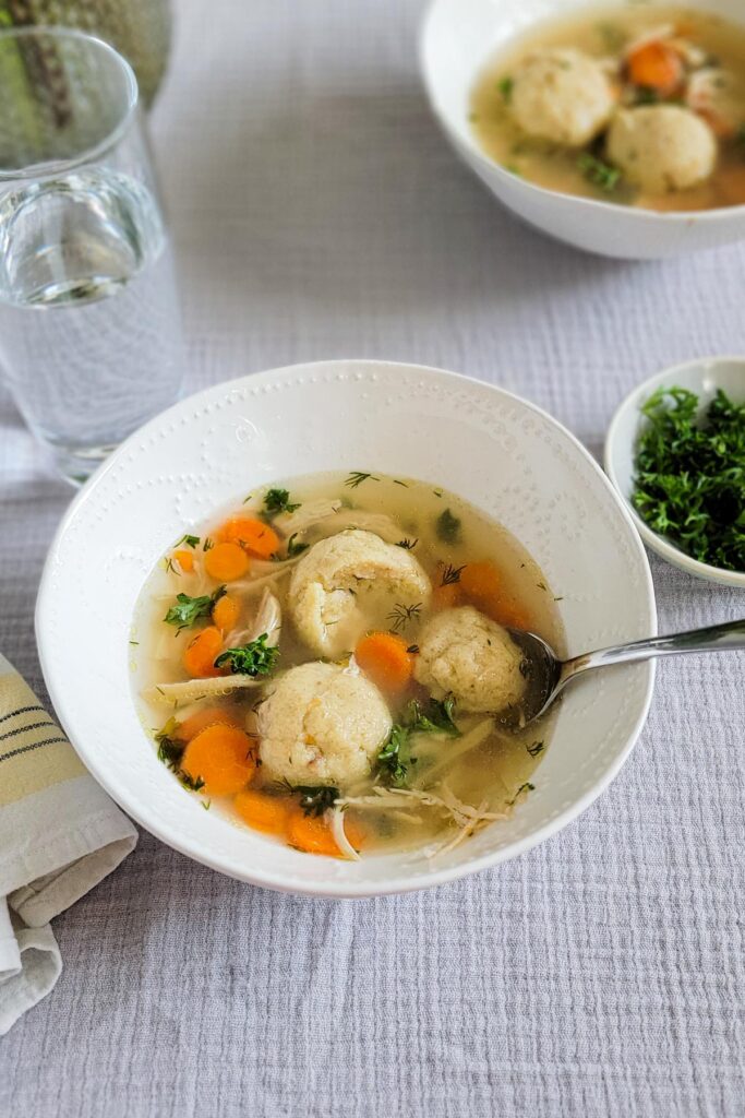 white bowl of matzo ball soup with carrots, parsley, dill and chicken. Spoon cutting into a matzo ball in the bowl. Glass of water and parsley garnish in the background.