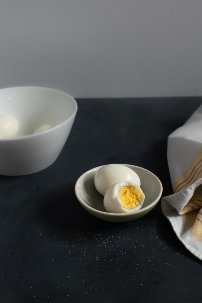 small green-toned dish with hard boiled eggs, one has a bite taken out. Salt sprinkled on black table surface. White napkin with yellow stripe on right side. White bowl with additional hard boiled eggs in background.