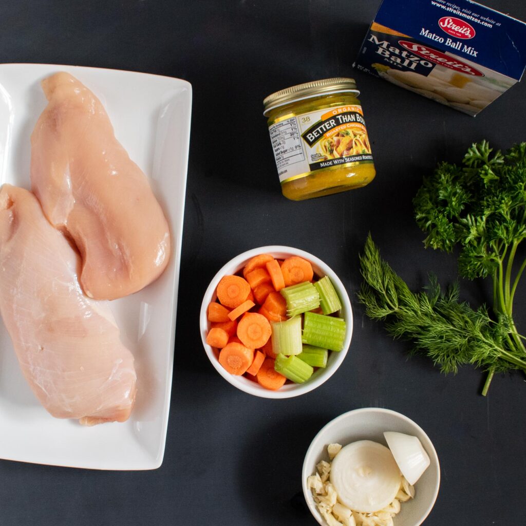 Display of soup ingredients. White rectangular plate with two raw chicken breasts, small white bowl of chopped carrots and celery, small white bowl of halved onion and chopped garlic, jar of chicken bullion base, box of streit's matzo ball mix, stems of parsley and dill. 