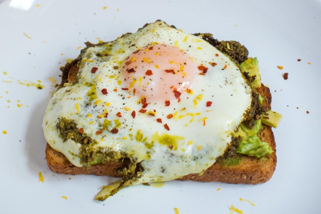 close up shot of sunny side up egg on a slice of toast with mashed avocado, on a white plate. Red pepper flakes and lemon zest sprinkled on top.