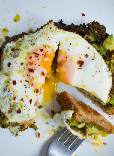 white plate with toast, mashed avocado and sunny side up egg that is cut to show runny yolk. Fork with a piece of the toast.