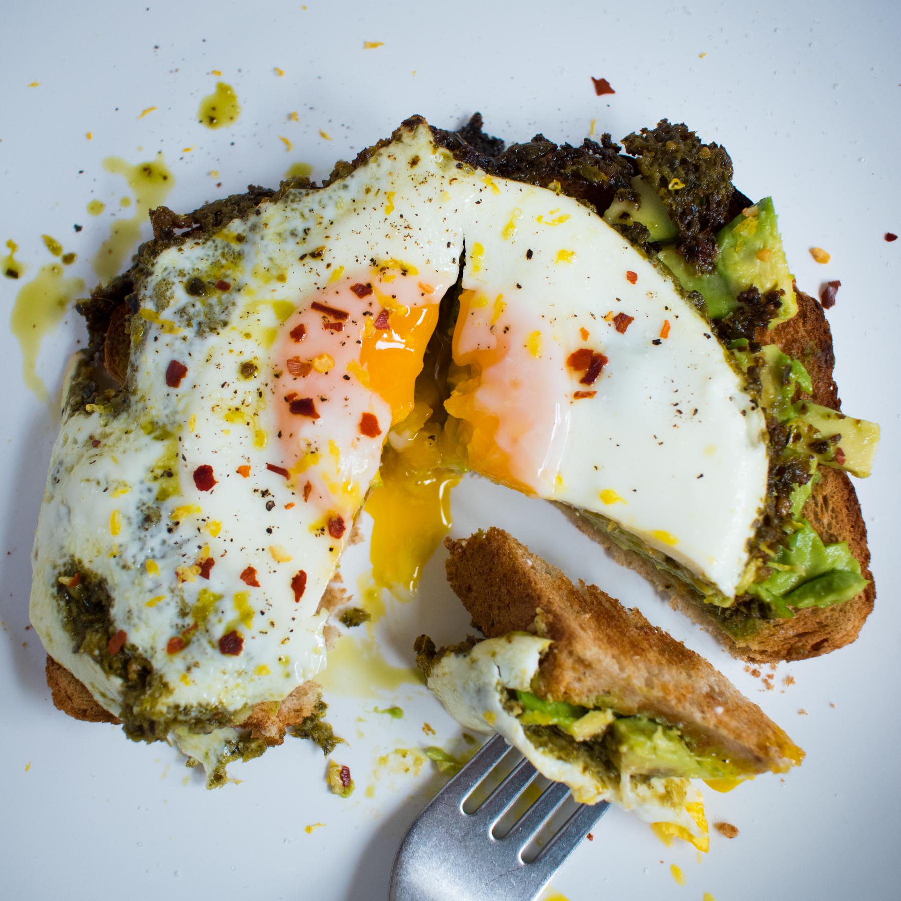 white plate with slice of toast topped with smashed avocado, pesto, fried egg that is broken into