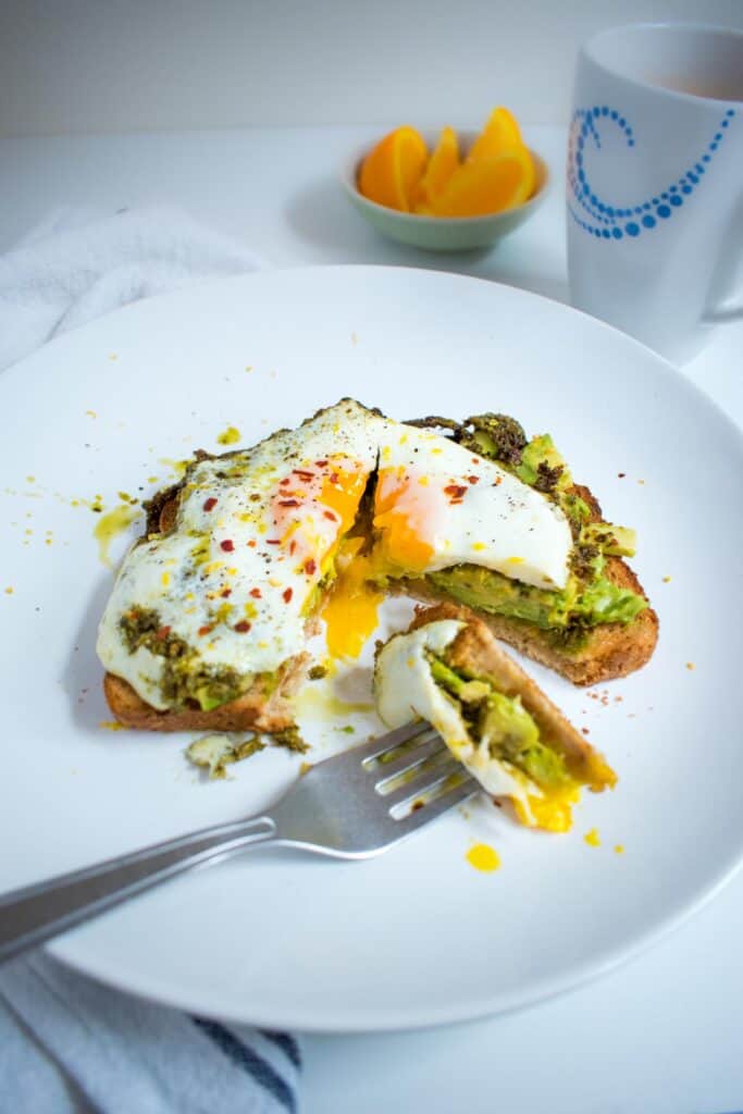 white plate with toast, mashed avocado and sunny side up egg that is cut to show runny yolk. Fork with a piece of the toast. Small dish of orange slices and mug of coffee in background.