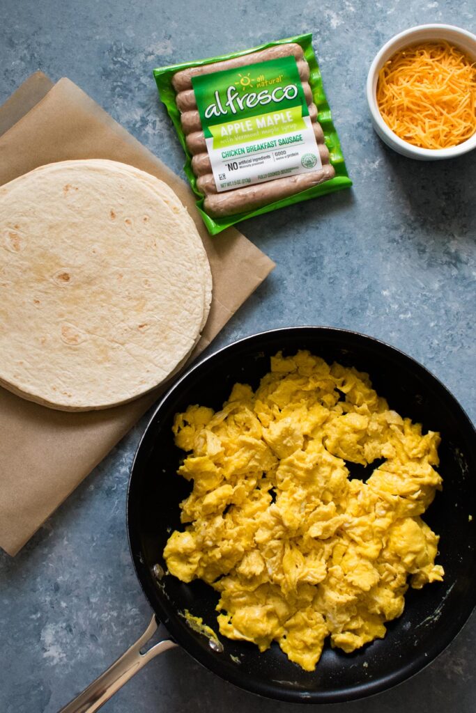 ingredients for burritos laid out; parchment paper with white tortillas, package of chicken breakfast sausages, white bowl of shredded cheddar, and black pan of scrambled eggs