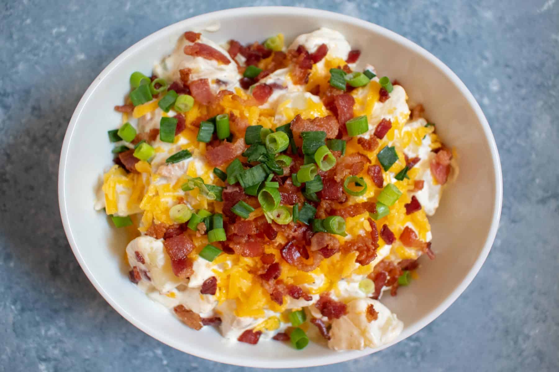 white bowl of potato salad topped with cheddar cheese green onions and bacon