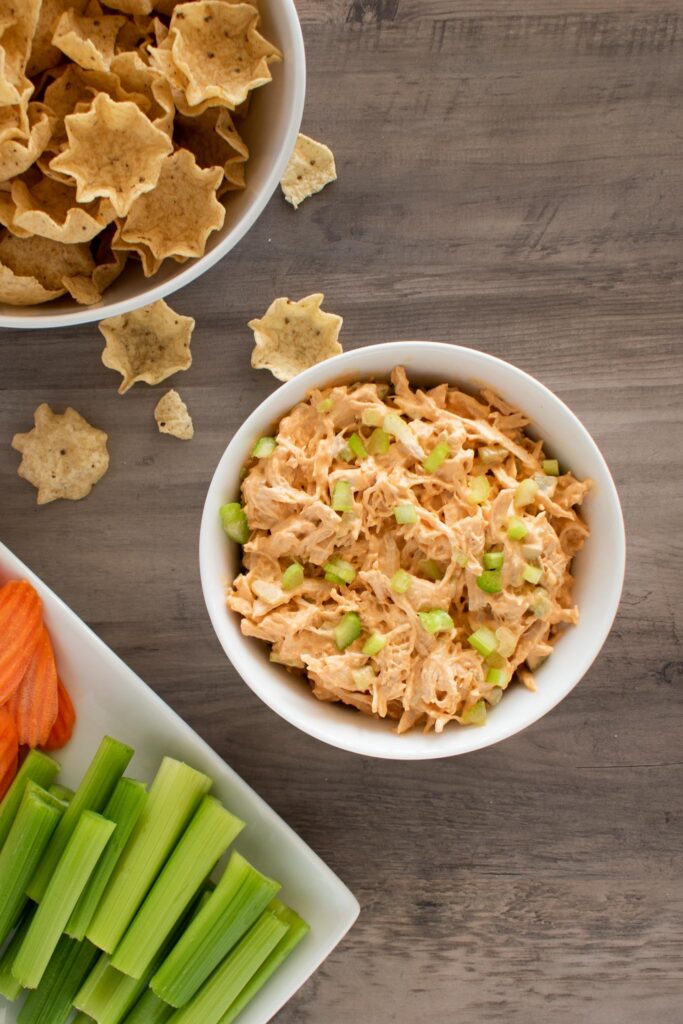 white bowl of buffalo chicken dip with a plate of sliced carrots and celery sticks and a bowl of tostitos scoops.