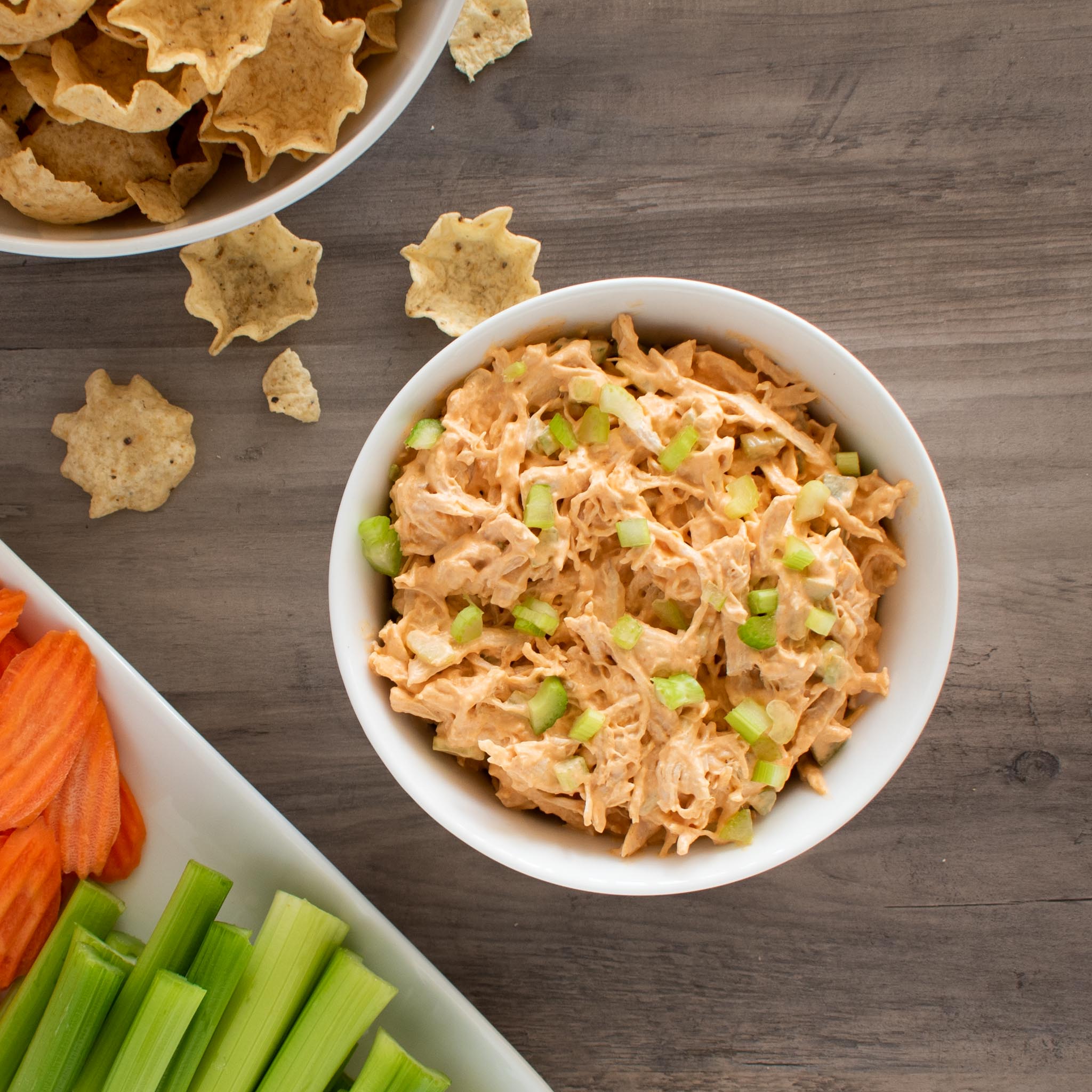 bowl of buffalo chicken dip, plate of celery sticks and carrot chips, bowl of tostitos chips.