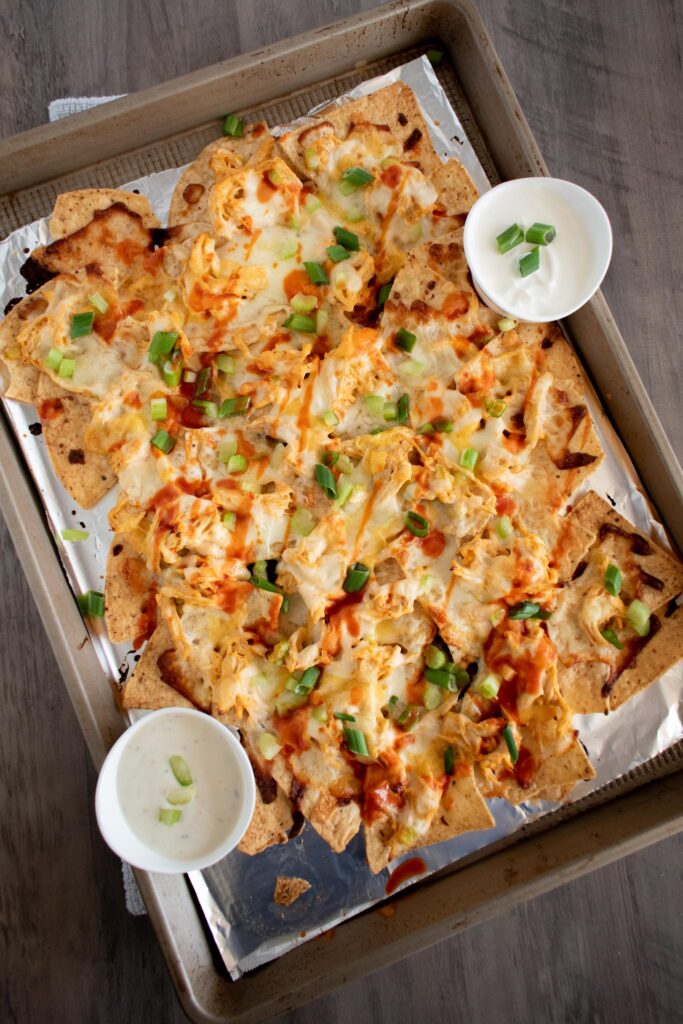 sheet pan of buffalo chicken nachos with two small dishes of sour cream and ranch.