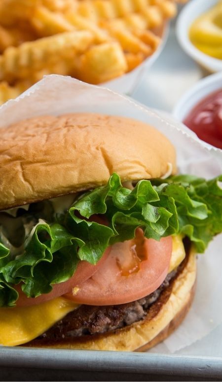 close up of a cheeseburger with lettuce and tomato, ketchup and mustard dishes and french fries in the background.