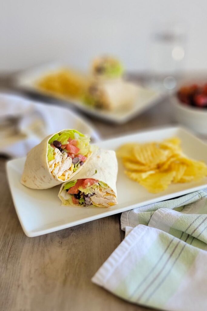 white plate with a chipotle ranch chicken wrap cut in half, side of potato chips, napkin.