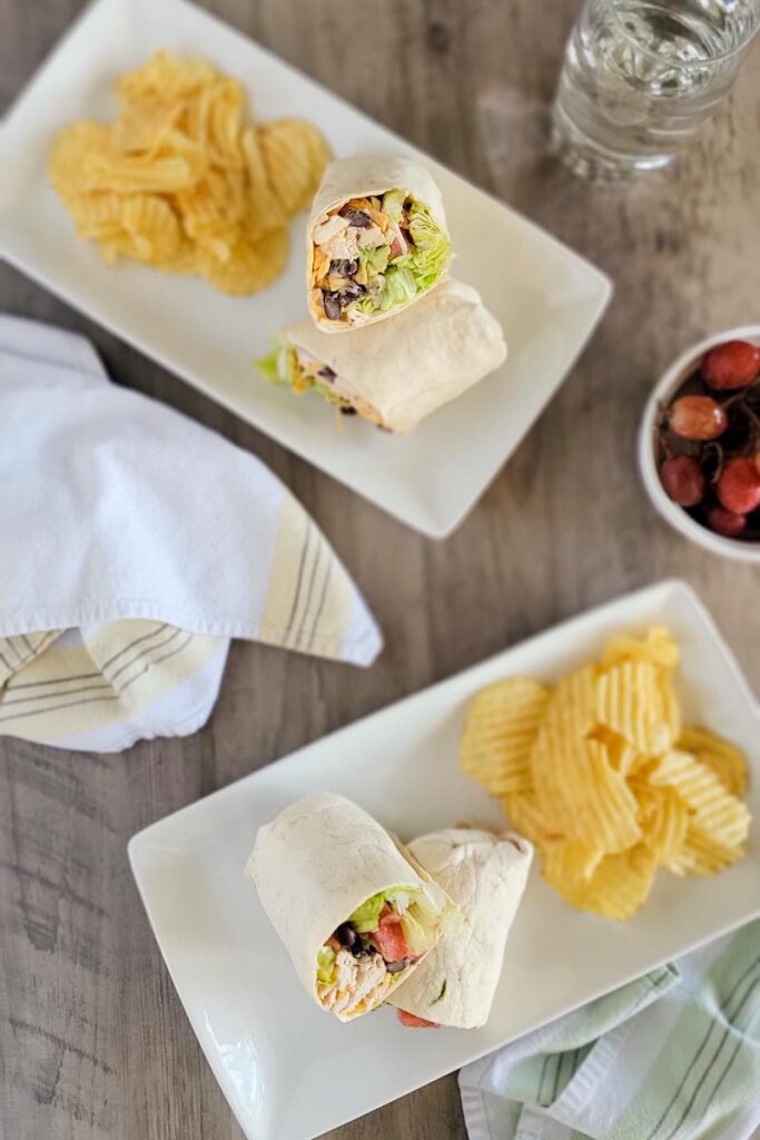 two white plates with wraps and potato chips. Side bowl of grapes. Napkins.