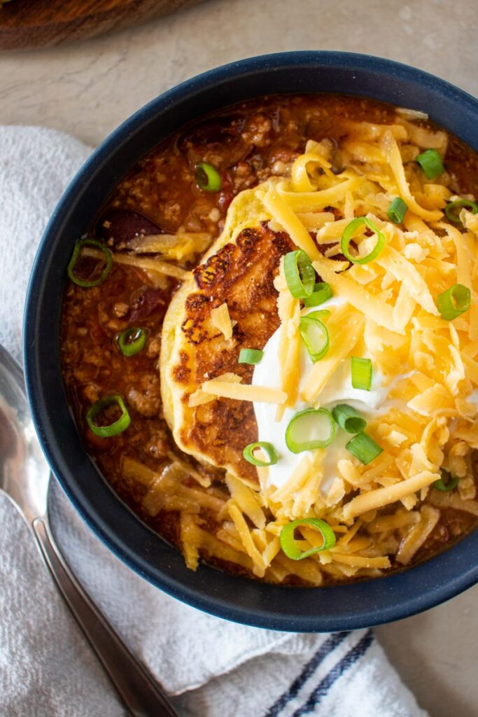 close up bowl of chili topped with corn cake, shredded cheddar, sour cream, and green onions.