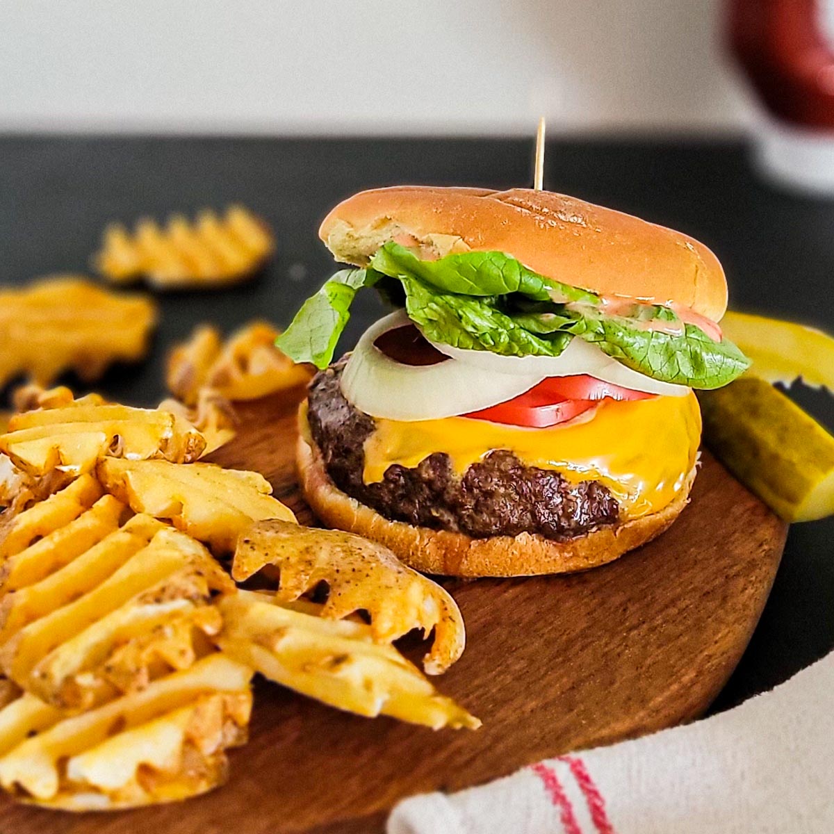 wood platter with a cheeseburger topped with melted cheese, lettuce, tomato, and onion. french fries and pickle spears on side.
