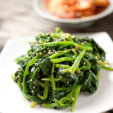 white plate of sauteed spinach with garlic, sprinkled with sesame seeds. Dish of food in background.