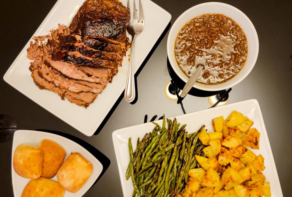white serving plates on a black glass table consisting of partially sliced beef brisket, onion au jus, roasted asparagus and potatoes, and bread rolls.